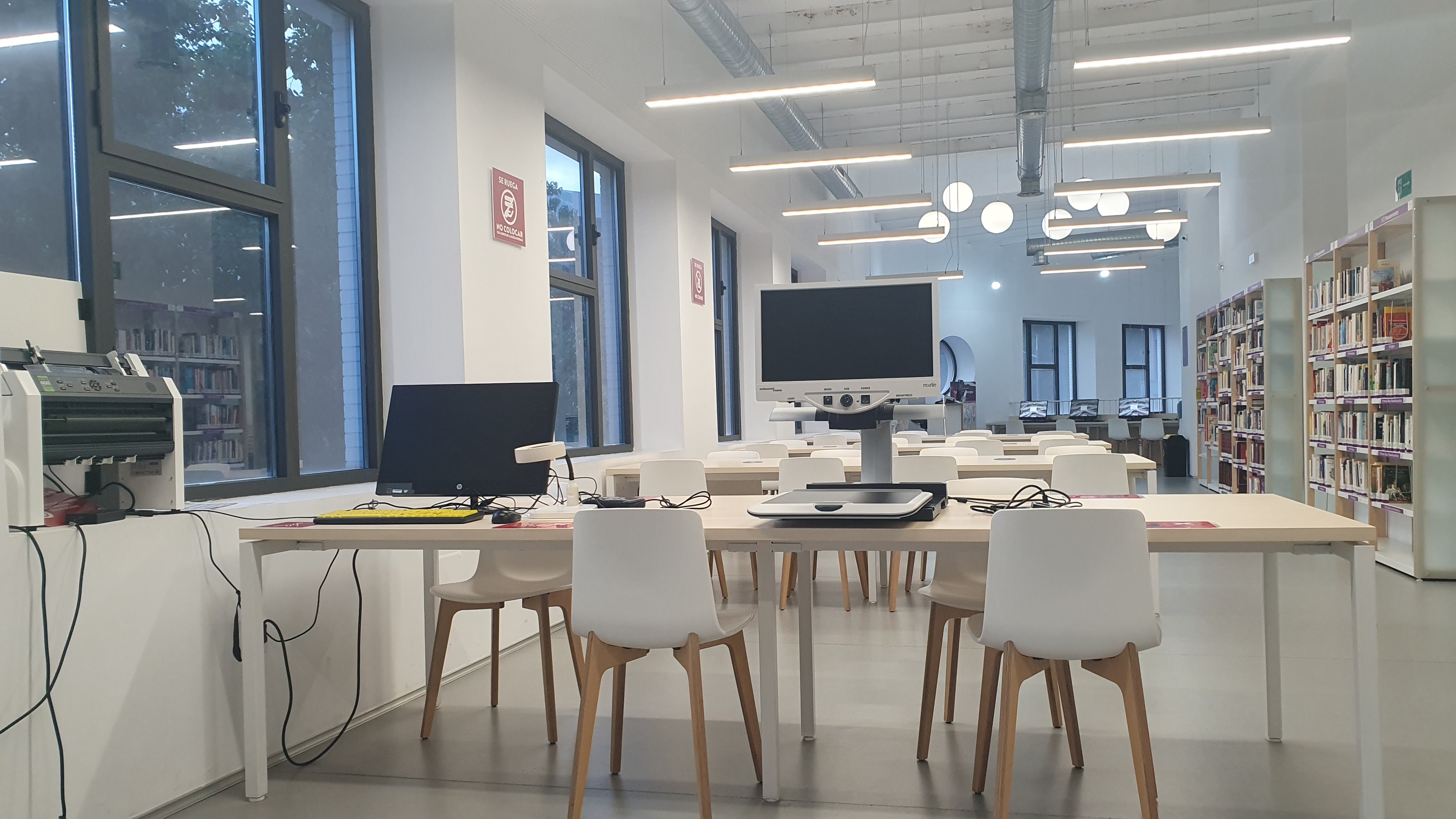 Sala de biblioteca donde se ven puestos para el trabajo de usuarios y una estantería llena de libros  