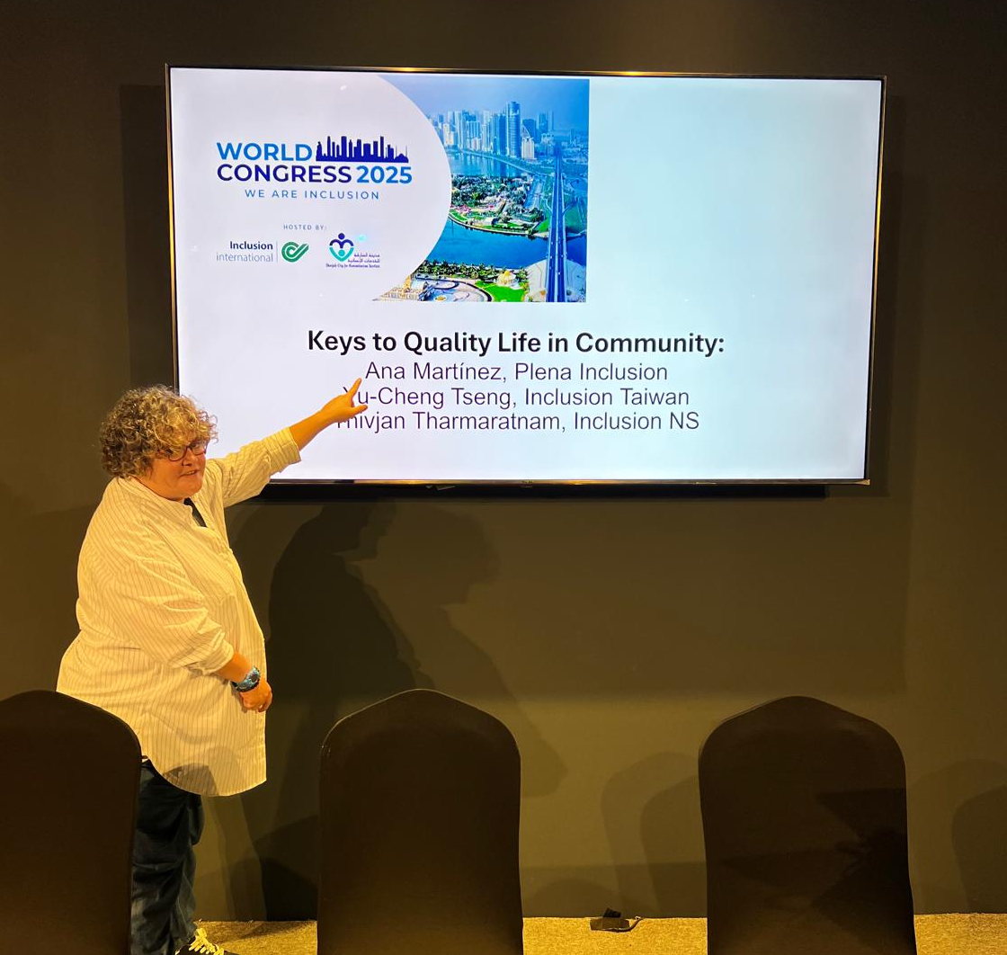 Ana señalando una pantalla con la presentación ‘Keys to Quality Life in Community’ durante el World Congress 2025, con logotipos de Inclusion International y vista panorámica de Dubái