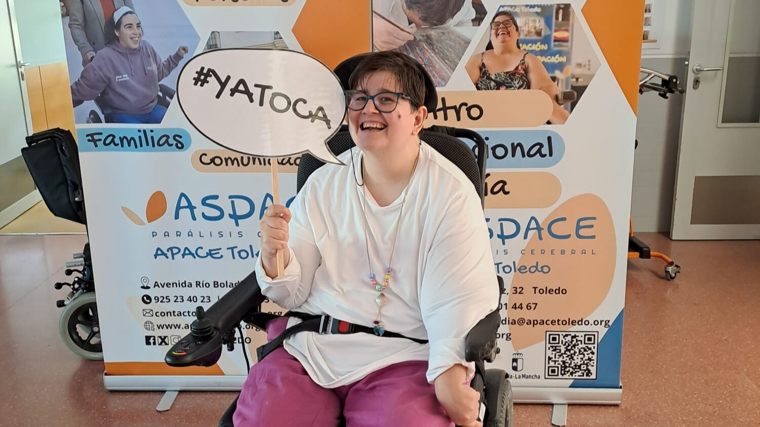 Ana Lidia en silla de ruedas sostiene un cartel con el lema '#YATOCA' frente a un panel informativo de ASPACE Toledo