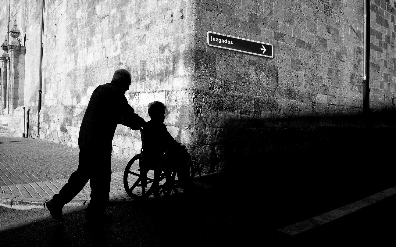 Fotografía en blanco y negro que muestra la silueta de una persona de pie y otra en silla de ruedas en un pasillo o túnel de ladrillo. Se ve una flecha que indica  'Juzgados'