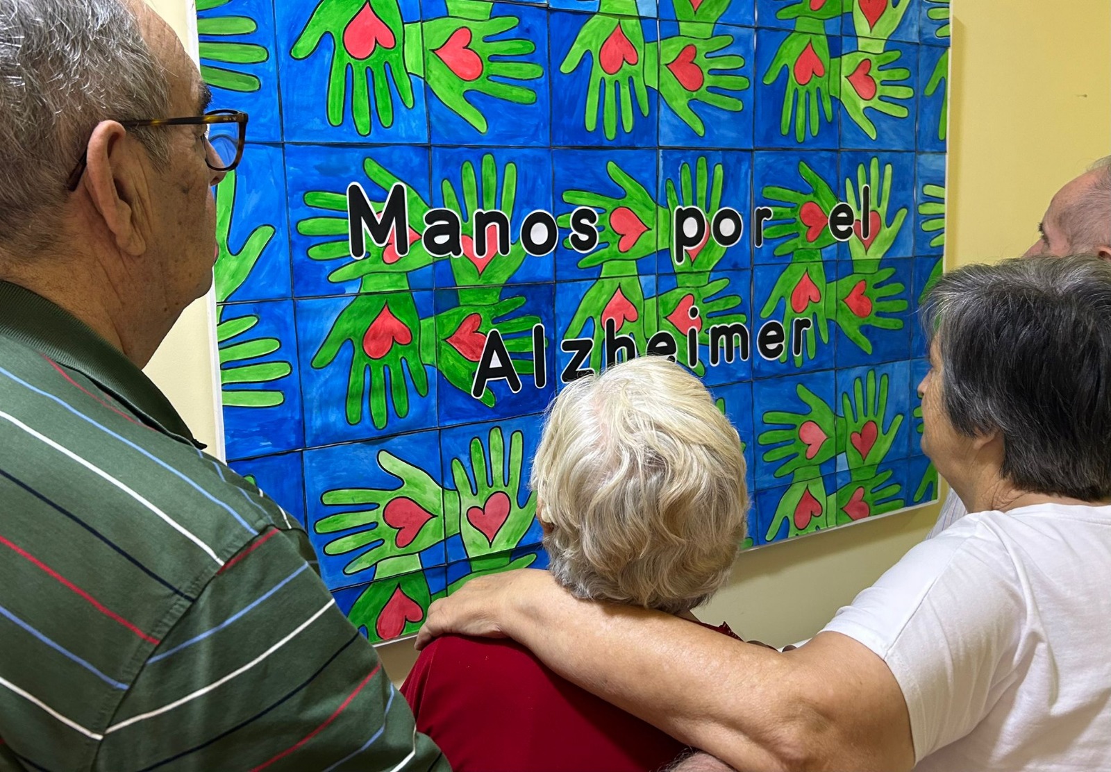 Imagen de personas mayores de espaldas. mirando un mural en el que se ven manos verdes y corazones rojos y se lee: “Manos por el alzheimer”