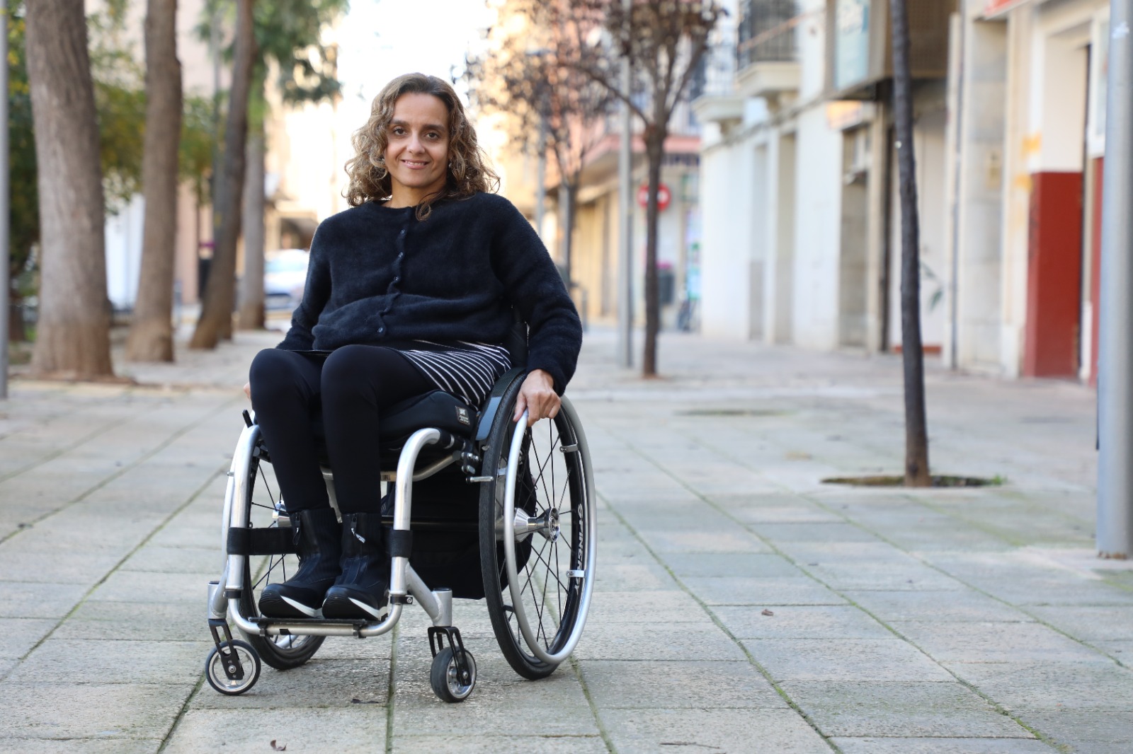 Fabiola en silla de ruedas sonriente sobre una acera, vestida con ropa oscura.