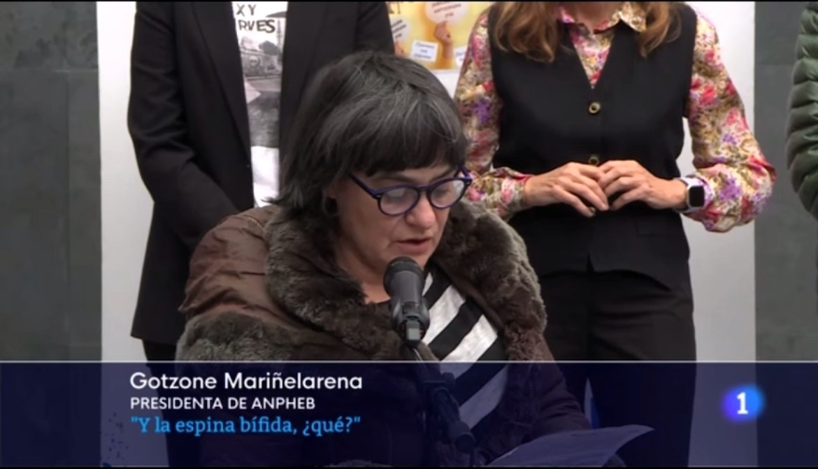 Foto de Gotzone hablando frente a un micrófono en la que se ve también un cartel que dice: “Gotzone Mariñelarena, presidenta de ANPHEB. ¿Y la espina bífida, qué?”.