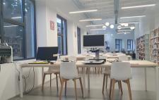 Sala de biblioteca donde se ven puestos para el trabajo de usuarios y una estantería llena de libros  
