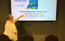 Ana señalando una pantalla con la presentación ‘Keys to Quality Life in Community’ durante el World Congress 2025, con logotipos de Inclusion International y vista panorámica de Dubái