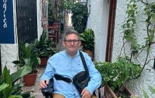 Imagen de José Luis en su silla de ruedas, en una calle estrecha llena de plantas