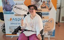 Ana Lidia en silla de ruedas sostiene un cartel con el lema '#YATOCA' frente a un panel informativo de ASPACE Toledo