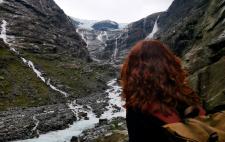 persona de espaldas con cabello rizado y rojizo que mira  hacia un paisaje de montañas parcialmente cubiertas de nieve