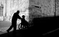 Fotografía en blanco y negro que muestra la silueta de una persona de pie y otra en silla de ruedas en un pasillo o túnel de ladrillo. Se ve una flecha que indica  'Juzgados'