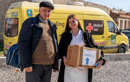Foto en la que se ve a una cooperante llevando una caja de farmacia, acompañada de un hombre  
