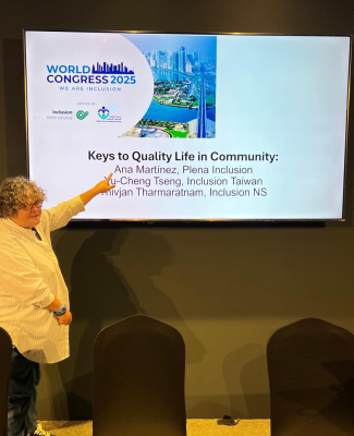 Ana señalando una pantalla con la presentación ‘Keys to Quality Life in Community’ durante el World Congress 2025, con logotipos de Inclusion International y vista panorámica de Dubái