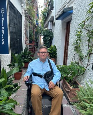 Imagen de José Luis en su silla de ruedas, en una calle estrecha llena de plantas