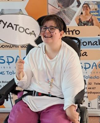 Ana Lidia en silla de ruedas sostiene un cartel con el lema '#YATOCA' frente a un panel informativo de ASPACE Toledo
