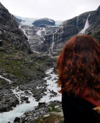 persona de espaldas con cabello rizado y rojizo que mira  hacia un paisaje de montañas parcialmente cubiertas de nieve