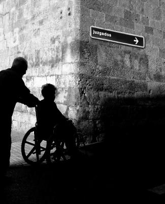 Fotografía en blanco y negro que muestra la silueta de una persona de pie y otra en silla de ruedas en un pasillo o túnel de ladrillo. Se ve una flecha que indica  'Juzgados'