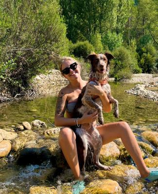 Imagen de Inmaculada sentada en rocas dentro de un arroyo, sosteniendo a un perro con collar rojo 